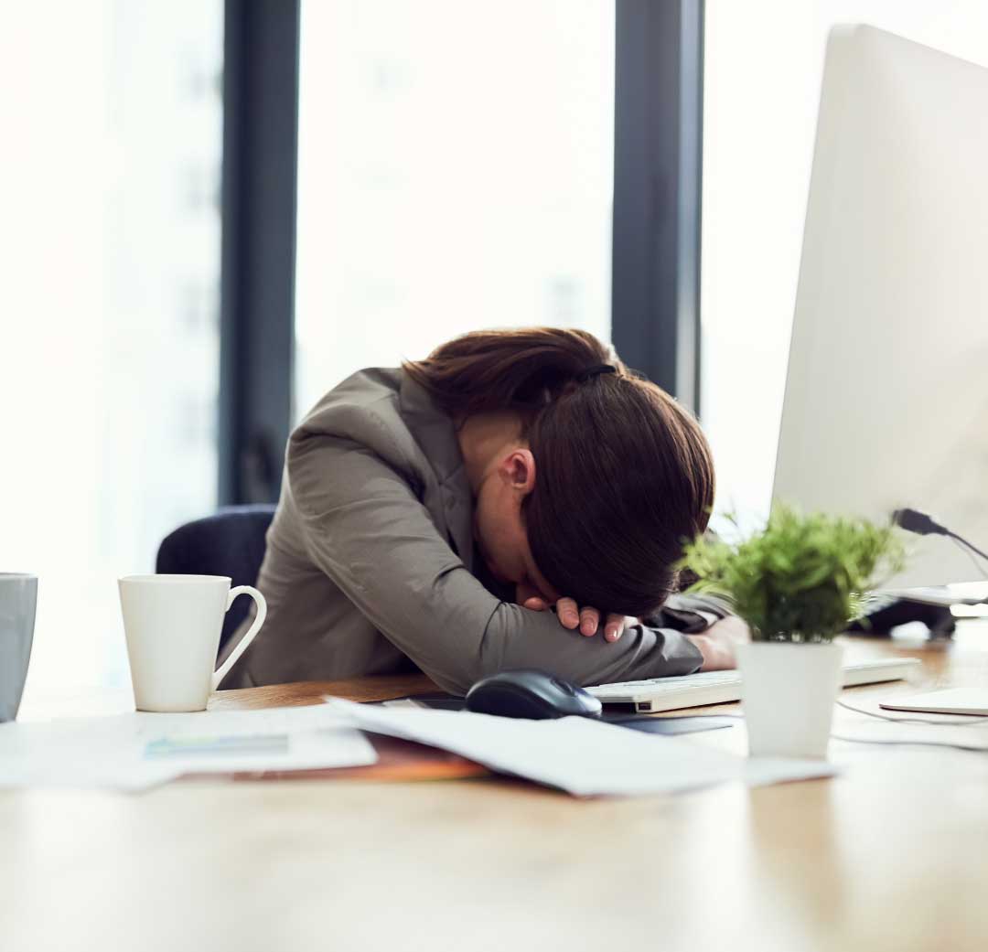 A woman at her desk with her head down on her folded arms, papers and an untouched coffee around her.