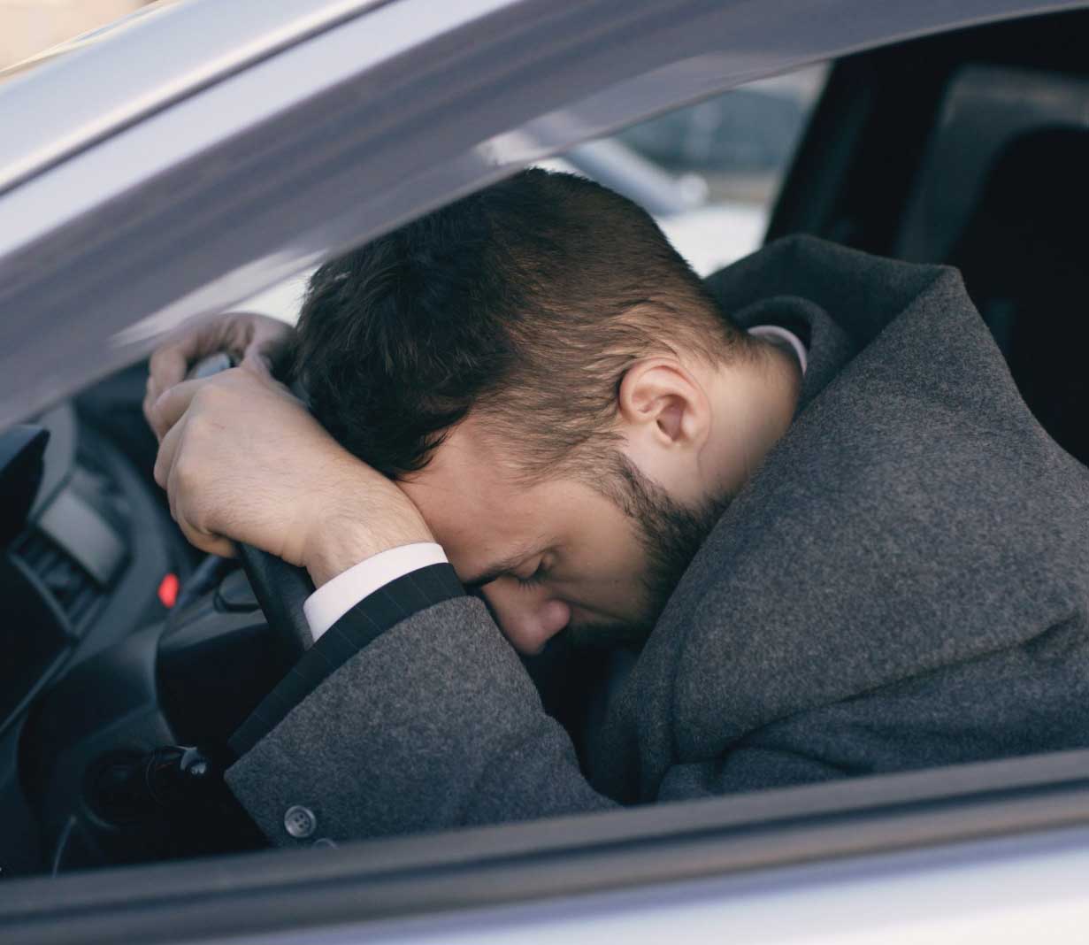 A man sitting in a parked car with his head resting on his arms on the steering wheel.