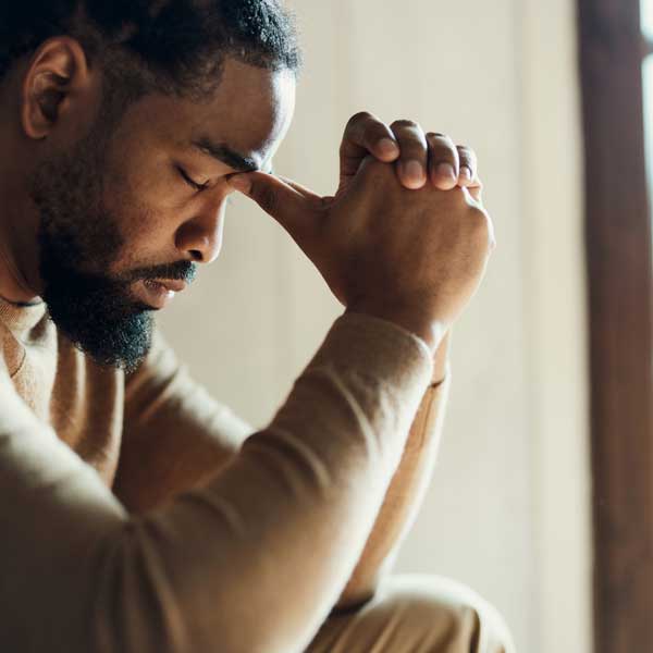 A man sitting with his hands clasped against his forehead, eyes closed.