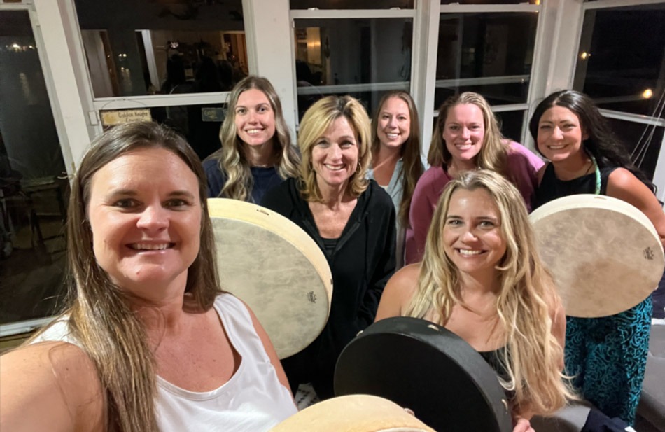 Seven women smiling together in the studio at night, several holding frame drums