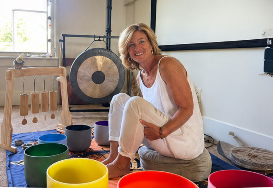 Dr. Tanya Lea smiling in the studio, seated among crystal singing bowls with a gong behind her
