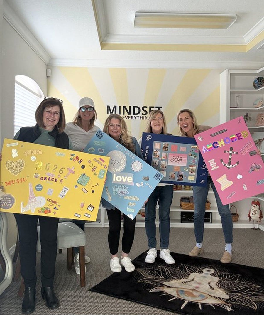Five women smiling in the studio, each holding up a colorful vision board, in front of a yellow sunburst mural